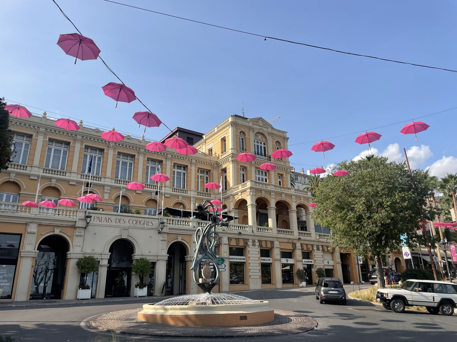 Grasse town square — pink umbrellas strung above, elegant ochre building, blue sky